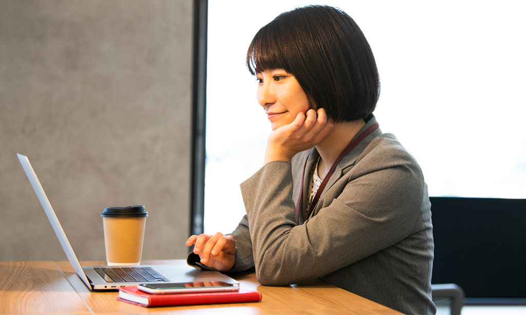 コーヒーを飲みながらノートPCで楽しげに作業する女性の画像